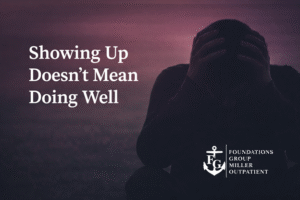Man sitting in a dimly lit living room, leaning forward with his head in his hands, conveying hidden emotional struggle; Foundations Group Miller Outpatient logo in corner.